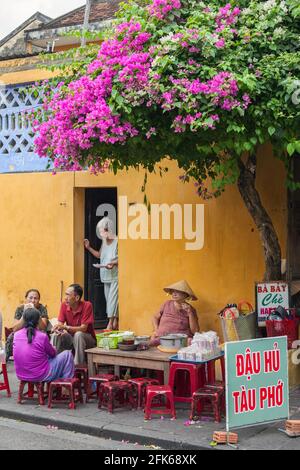 Vietnamese female selling street food at roadside cafe beneath bougainvillea flowers, Hoi An, Vietnam Stock Photo