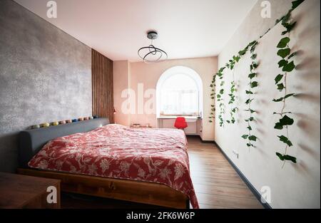 Stylish interior of bedroom with gray and pink color of walls. King-size bed covered red sheet, green plant hanging on pink wall and cozy workplace under arch near window. Stock Photo