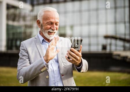 Happy senior businessman is relaxing after work in front of company building. He is messaging on smartphone. Stock Photo