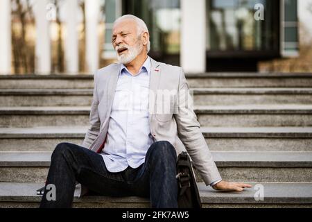 Senior businessman is tired of work. He is sitting on stairs in front of company building. Stock Photo