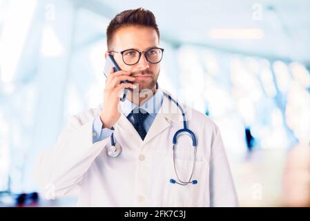 Male doctor using his mobile phone and talking with somebody while standing at the hospital’s corridor. Stock Photo