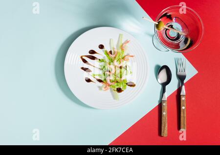 Spring seafood salat of shrimps, greens, celery, balsamic sauce with martini cocktail in white plate on red and minty color background with shadow in Stock Photo