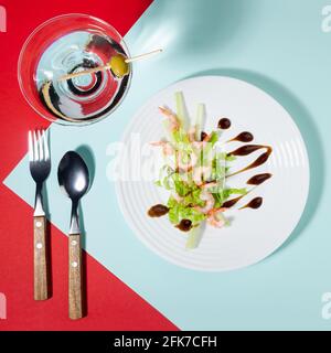 Fresh summer salad of shrimps, greens, celery, teriyaki sauce with cutlery, with martini glass on red and minty color with shadow in sunlight, square. Stock Photo