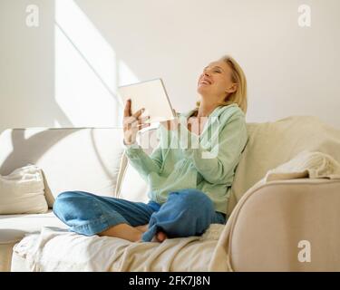 Overjoyed laughing senior woman in casual wear watching funny video or movie on digital tablet while sitting on sofa and relaxing at home, happy elderly female enjoying free time and retired life Stock Photo
