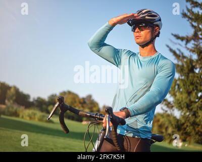 Professional male cyclist wearing sportswear, protective helmet and glasses holding hand on forehead while looking away, standing with a bike in park Stock Photo