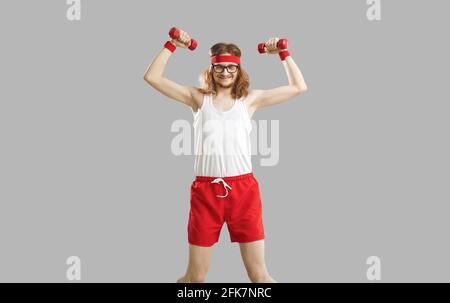 Funny skinny man with dumbbells exercising weak muscles isolated on gray background Stock Photo