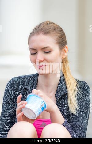 Beautiful lovely young blonde hair girl smiling and eating yogurt in the city, vertical Stock Photo