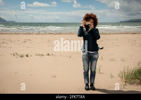 Beautiful young woman with curly red hair holding an wooden toy photo camera on empty sandy beach background. Fun photography concept. Stock Photo