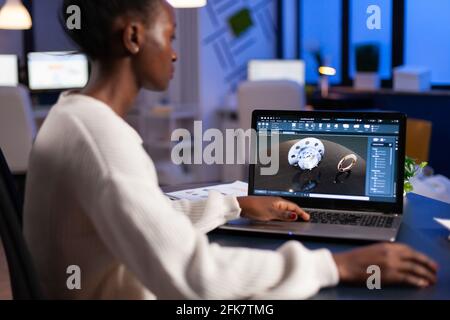 Tired african designer engineer analyzing new prototype of 3D model of industry product, working overtime. Industrial female worker studying gears idea on laptop showing cad software on device display. Stock Photo