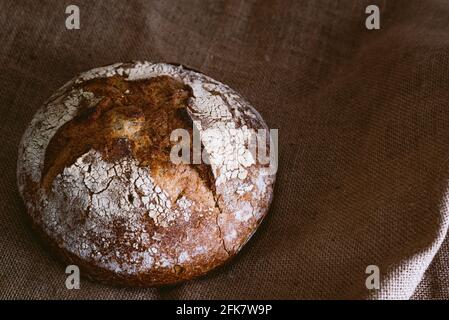 fresh round loaf of healthy whole grain bread on brown jute burlap fabric background with white flour on top. top view of homemade bread on hessian fa Stock Photo