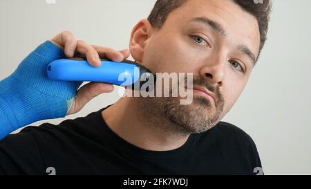 Male hand in blue waterproof bandage shaves stubble with a razor on a bright background. Man with hand wrapped in fiberglass plaster cast shaves his cheek with an electric safety razor Stock Photo