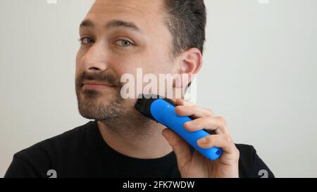 Male hand smoothly shaves stubble with shaving machine on bright background. Handsome middle-aged man shaves his cheek with an electric safety razor. Shaving the beard on the face Stock Photo