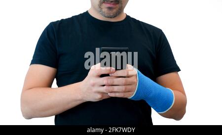 Unrecognizable middle-aged bearded man in a black T-shirt with a broken wrist uses phone against a white background. Male hand, wrapped in a fiberglass plaster cast, holds mobile phone Stock Photo