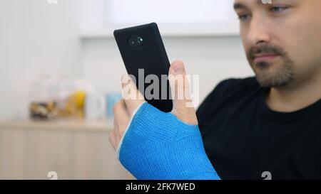 Handsome middle-aged man in black t-shirt with broken wrist surfs the internet at home. Male hand, wrapped in a fiberglass plaster cast, uses a touchscreen on a mobile phone Stock Photo