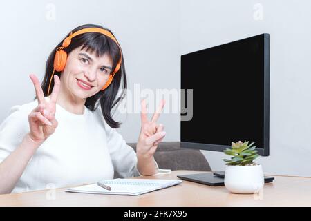 Smiling business woman in headphones looks at you, sitting at the table in front of the monitor with a blank black screen and shows with her hands a s Stock Photo