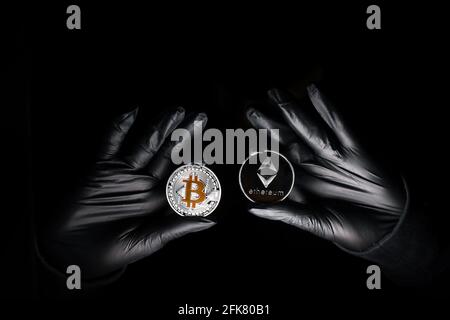 Hands in a black gloves holding bitcoin and Ethereum coin against black background. International hacker is trying to steel your crypto currencys. Blockchain security. Stock Photo