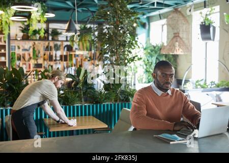 Portrait of African-American businessman using laptop while working in eco friendly cafe interior decorated with fresh green plants, copy space Stock Photo