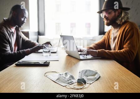 Portrait of two contemporary men using laptops while working at cafe table with focus on two face masks in foreground, covid concept, copy space Stock Photo