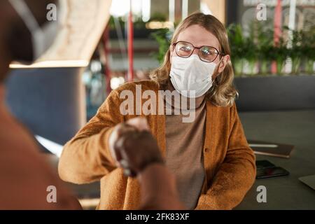 Portrait of long haired man wearing mask and bumping fist with colleague during con tactless greeting in cafe or office, covid concept Stock Photo