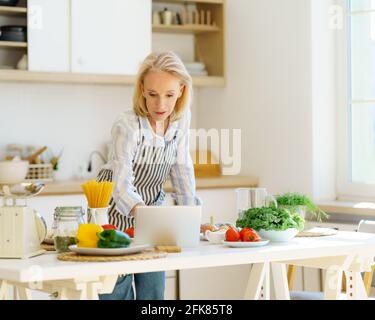Senior 60s woman in apron learning how to cook, watching online culinary class or video recipe on digital tablet, standing in kitchen. Eldrely female exploring new cuisines while cooking at home Stock Photo