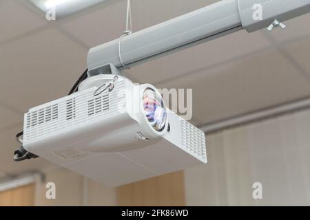 A white video projector hangs from the ceiling with an adjustable arm. Against the background of light ceiling panels. Round objective lens with abstract pattern. Stock Photo