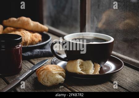 Hot cup of coffee with croissants on a rustic wooden table next to window Stock Photo