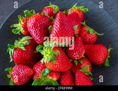Overhead and closeup view of a dark plate with fresh strawberries with stem and leaves on black table background Stock Photo