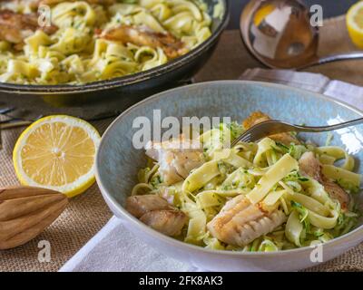 Italian creamy pasta dish with tagliatelle, zucchini and fried white fish fillet served on a plate with pan in the background on kitchen table Stock Photo