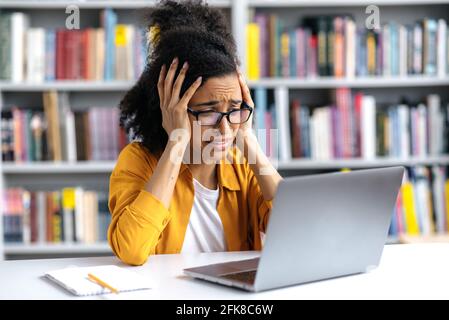 Upset shocked young woman, african american student girl, wearing glasses, sits at a table in a library, clasped her head in horror, looking at laptop screen, emotional face expression Stock Photo