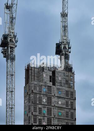 A pair of tower cranes, working to construct a central building core bearing Android logos, against an overcast grey sky. Stock Photo