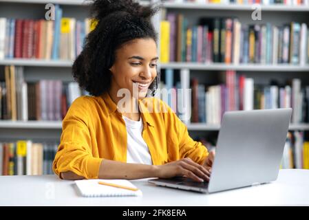 Happy attractive african american girl in stylish clothes, female student sits in the university library at the desk, preparing for the exam or lesson, browsing internet, searching information, smiles Stock Photo