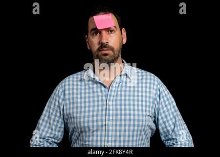 Bearded man in a white and blue plaid shirt holding a paper stuck to his forehead isolated on black studio background. Fun concept Stock Photo