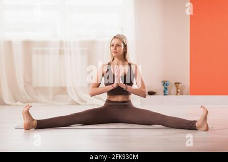 Woman in sportswear practicing yoga, sitting on a mat and doing cross twine in the studio with namaste. Stock Photo