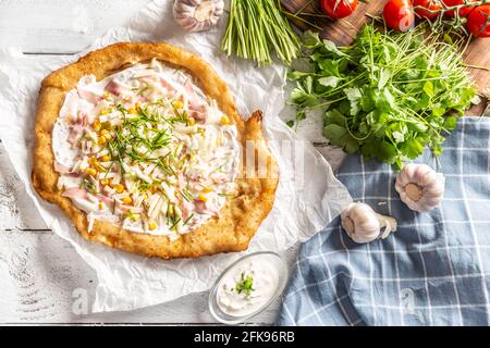 Tasty hungarian langos served with sour creme, ham, cheese, herbs and vegetables in the background. Stock Photo