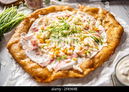 Tasty crusty langos served with sour cream, grated cheese and ham and corn with herbs. Stock Photo