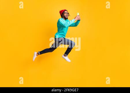 Full size profile photo of brunette optimistic guy jump run take photo wear cap spectacles pullover jeans isolated on yellow background Stock Photo