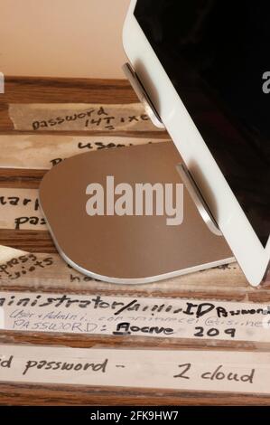 Passwords for computer tablet are written down and visible on desk top. The tablet computer is sitting upright on a holder and covering some writing. Stock Photo