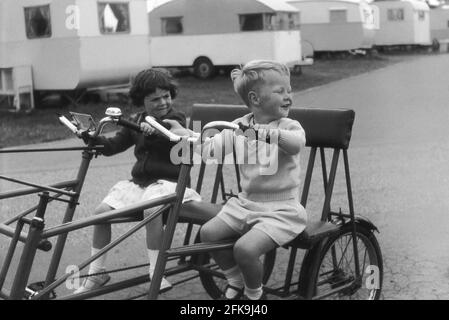 1964, historical, outside on a path at a holiday caravan park, a young boy with his little sister, sitting on a large metal framed 'holiday camp' twin or two seater bicycle, Suffolk, England, UK. These double seater bicycles were great fun and a good way of moving around the site on the wide paths. Stock Photo