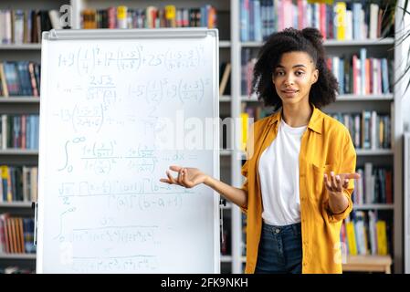 Portrait friendly young woman, african american female teacher, standing near whiteboard, in stylish clothes, conducts lecture or webinar by video call, gesturing with her hands, looks at camera Stock Photo