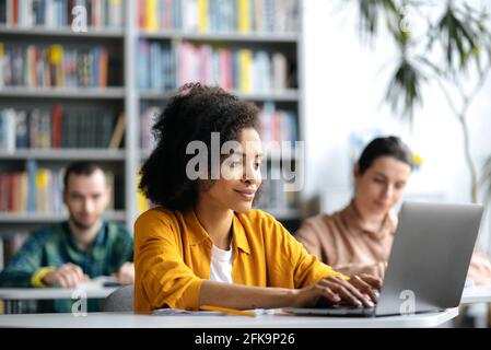 Focused smart attractive african american female student sitting at table in campus library with laptop while preparing for lessons or exam, browsing internet, two blurry students in the background Stock Photo