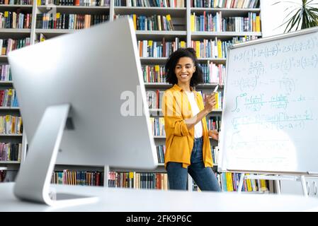 Joyful african american female coach, standing near whiteboard, in casual clothes, conducts an online webinar, communicating with students by conference call, looking at computer screen, smiling Stock Photo