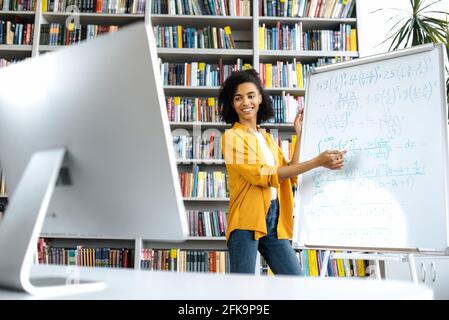 Stylish African American female coach, stands at whiteboard, in casual clothes, uses computer, leads an online lesson, communicates with students via video conference, looks at screen,distant learning Stock Photo