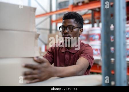 Busy young African-American warehouse employee in eyeglasses standing at metal frame and adjusting boxes Stock Photo