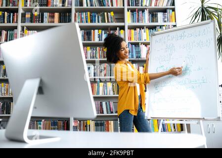 Stylish African American female coach, stands at whiteboard, in casual clothes, uses computer, leads an online lesson, communicates with students via video conference, looks at screen,distant learning Stock Photo