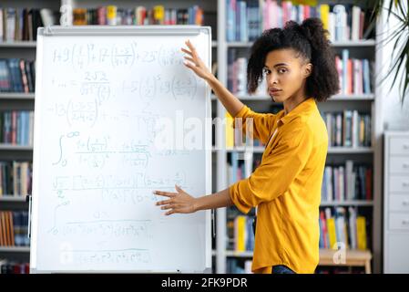 Serious young woman, mixed race teacher standing near whiteboard, wearing casual clothes, explains theme for students by video conference, gesturing with hands, looking at camera Stock Photo