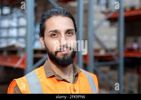 Portrait of smiling handsome young mixed race worker in orange vest standing against metal shelves at storehouse Stock Photo