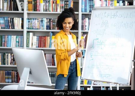 Young attractive smart African American woman, teacher, stands at the blackboard, uses a computer, conducts an online seminar, communicates with students via video call, gestures with her hand, smiles Stock Photo