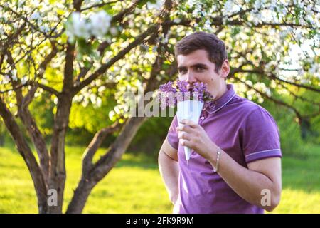 Defocus portrait of handsome caucasian man with cone flowers in lilac garden Funny enjoyment young brunette guy smelling fresh flowers and looking at Stock Photo