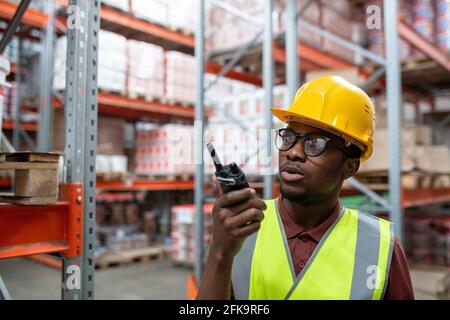 Serious young African-American storeroom manager in yellow hardhat and glasses using radio equipment while transmitting message to colleague Stock Photo