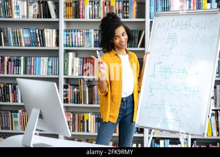 Happy smart confident beautiful African American female teacher in stylish wear, stands near the whiteboard, leads an online lesson, communicates with students through a video conference, by computer Stock Photo
